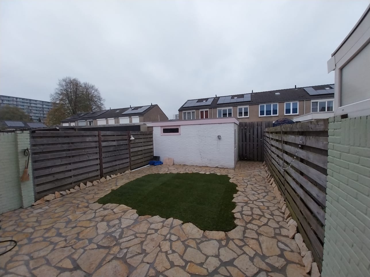 Ogród z nową trawą i ścieżką z kamieni, otoczony drewnianym ogrodzeniem. W tle biały budynek gospodarczy i zabudowa szeregowa z panelami słonecznymi.