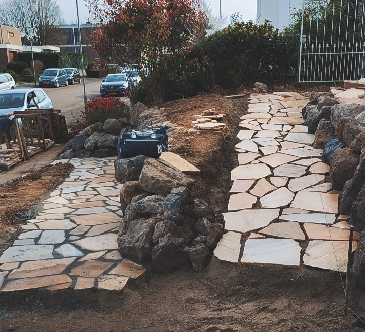 Ścieżka z nieregularnych płyt kamiennych, obłożona kamieniami, prowadząca przez ogród. W tle widoczne budynki i samochody.