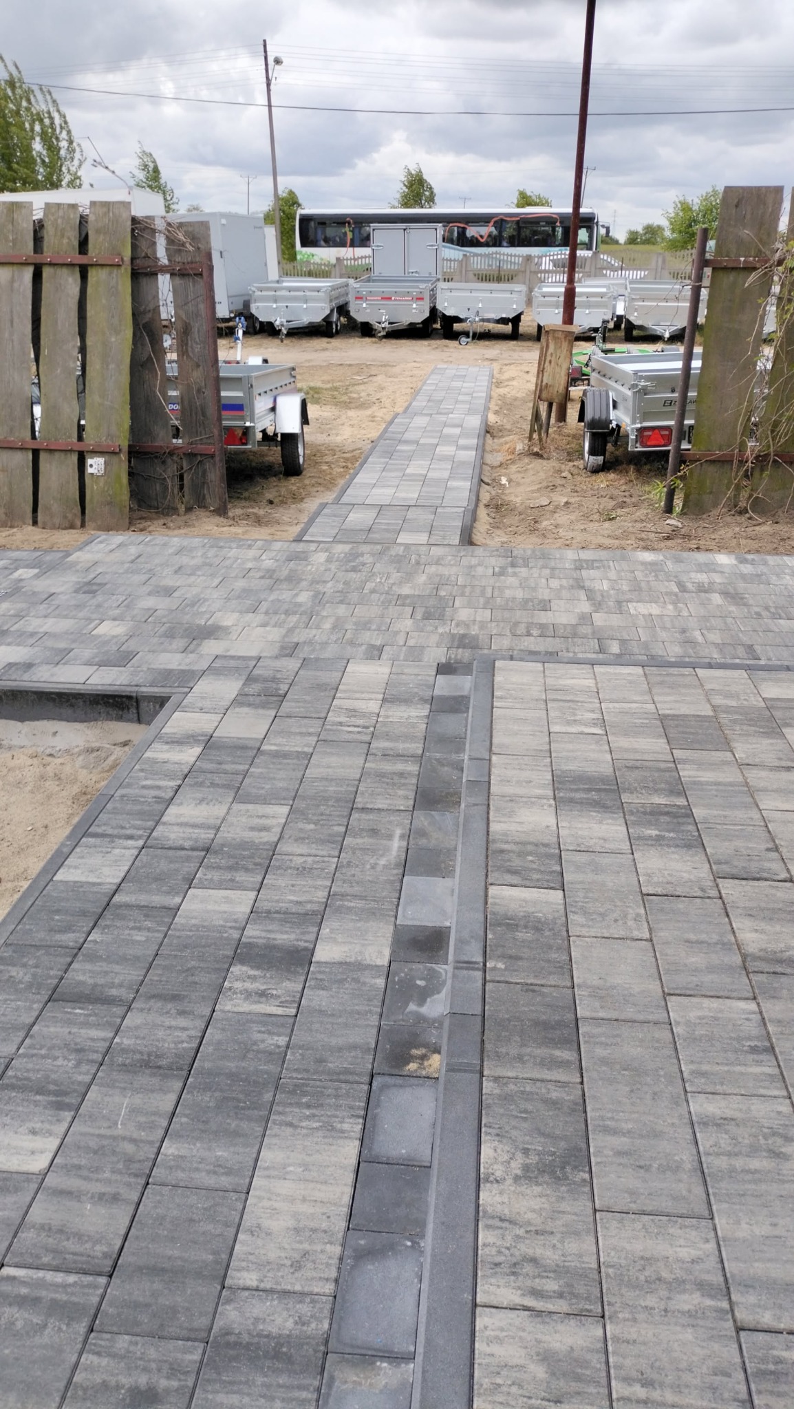 Szara kostka brukowa ułożona w geometryczny wzór, tworząca ścieżkę. W tle przyczepy i autobus, a po bokach drewniane elementy ogrodzenia.