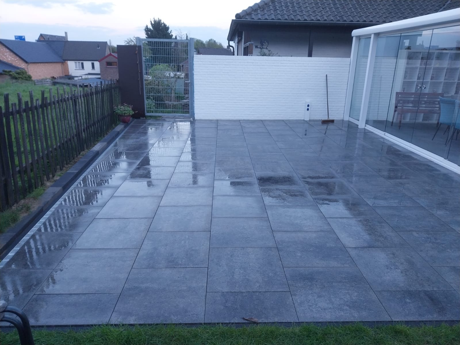 Nowoczesny taras z szarych, prostokątnych płyt. Widoczne ogrodzenie z metalowej siatki, drewniany płot i biała ściana z cegły. Powierzchnia płyt mokra, odbija światło.