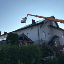 Tomasz Szatkowski - Pracownik w podnośniku koszowym czyści panele słoneczne na dachu dwupiętrowego domu. Widok z dołu, zieleń w tle.