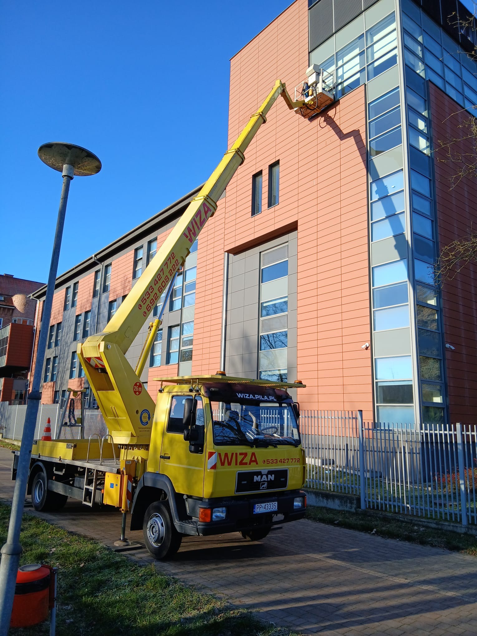 Żółty podnośnik koszowy firmy WIZA na tle budynku z pomarańczową elewacją i szklanymi oknami. Pracownik w koszu na wysokości wykonuje prace.
