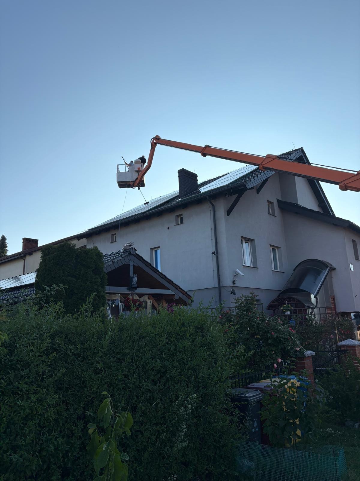 Pracownik w podnośniku koszowym czyści panele słoneczne na dachu dwupiętrowego domu. Widok z dołu, zieleń w tle.