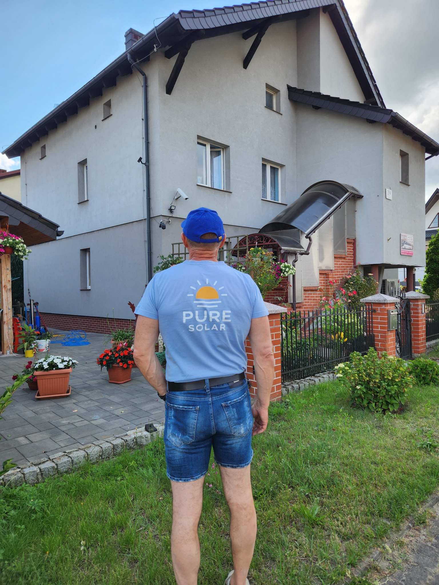 Mężczyzna w koszulce Pure Solar stoi tyłem na tle domu z kostki brukowej i trawnika. Widoczne kwiaty w donicach i elementy instalacji na elewacji.