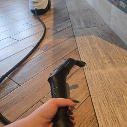 Sylwia Szwed - Czyszczenie fug parownicą Karcher na podłodze z płytek imitujących drewno. W tle ściana z dekoracyjnego kamienia i zasłona. Widoczna dłoń z zielonymi paznokciami.