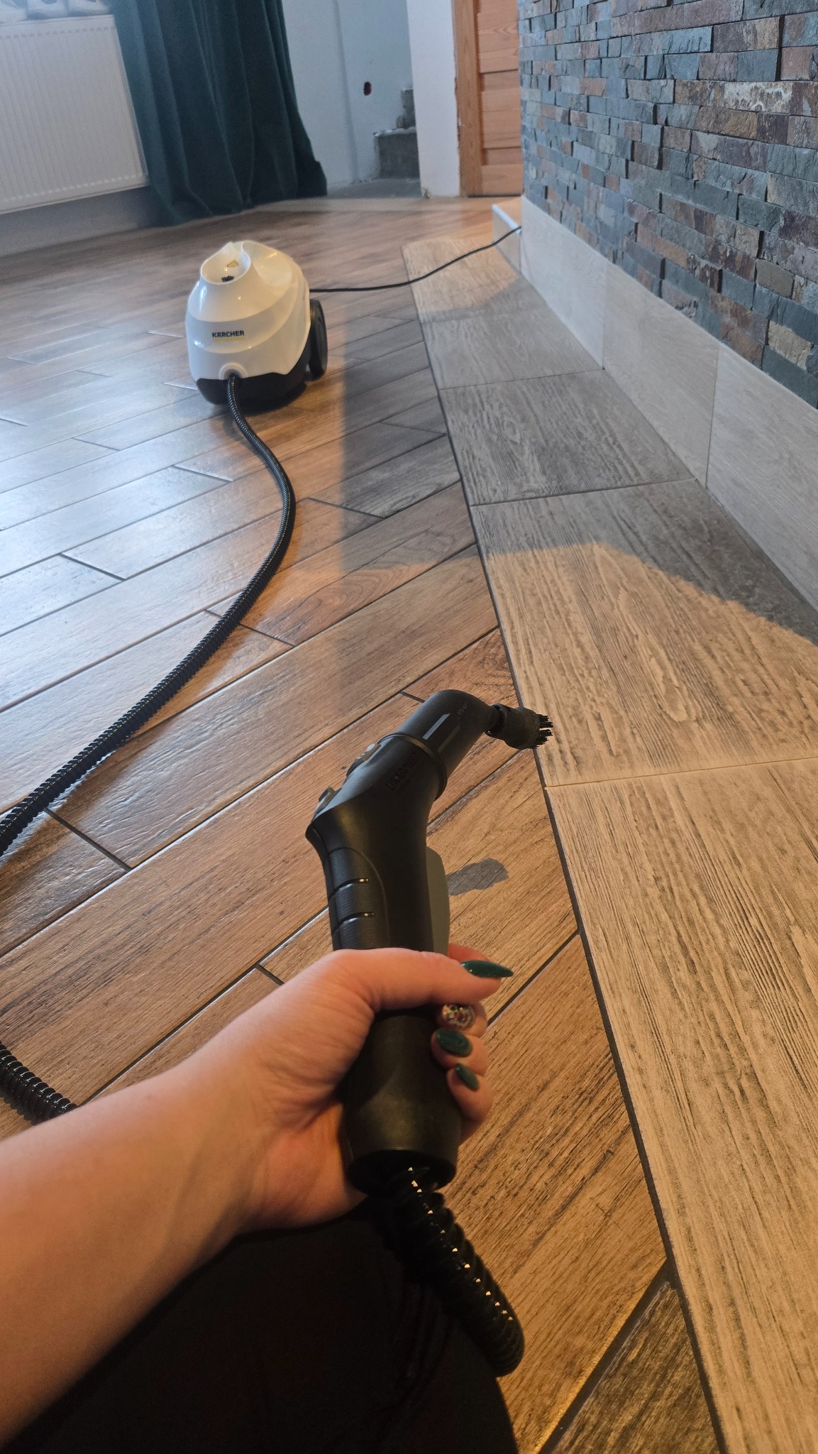 Czyszczenie fug parownicą Karcher na podłodze z płytek imitujących drewno. W tle ściana z dekoracyjnego kamienia i zasłona. Widoczna dłoń z zielonymi paznokciami.
