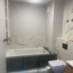 WOJCIECH BIERNACKI - Nowoczesna łazienka po remoncie: biała wanna z szarą obudową, marmurowe płytki, podświetlenie LED pod wanną, wisząca toaleta i minimalistyczne lampy.