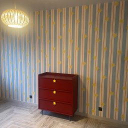 WOJCIECH BIERNACKI - Pokój dziecięcy po odświeżeniu: wzorzysta tapeta w paski i gwiazdki, czerwona komoda z żółtymi uchwytami w kształcie gwiazd, parkiet ułożony w jodełkę, wiklinowa lampa.