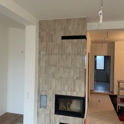 WOJCIECH BIERNACKI - Obudowa kominka z beżowych, błyszczących płytek ułożonych pionowo, z czarnymi akcentami. Widoczne fragmenty podłogi i sufitu w trakcie prac wykończeniowych.