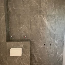 WOJCIECH BIERNACKI - Wykończona ściana łazienki z szarych, marmurkowych płytek, widoczne przyłącza wodne i otwór rewizyjny. Minimalistyczny design, nowoczesne wykończenie.