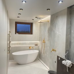 WOJCIECH BIERNACKI - Nowoczesna łazienka w trakcie wykańczania: biała wanna wolnostojąca, złota armatura, podświetlenie LED i fragment ściany w stanie surowym. Widoczne rury i instalacje.