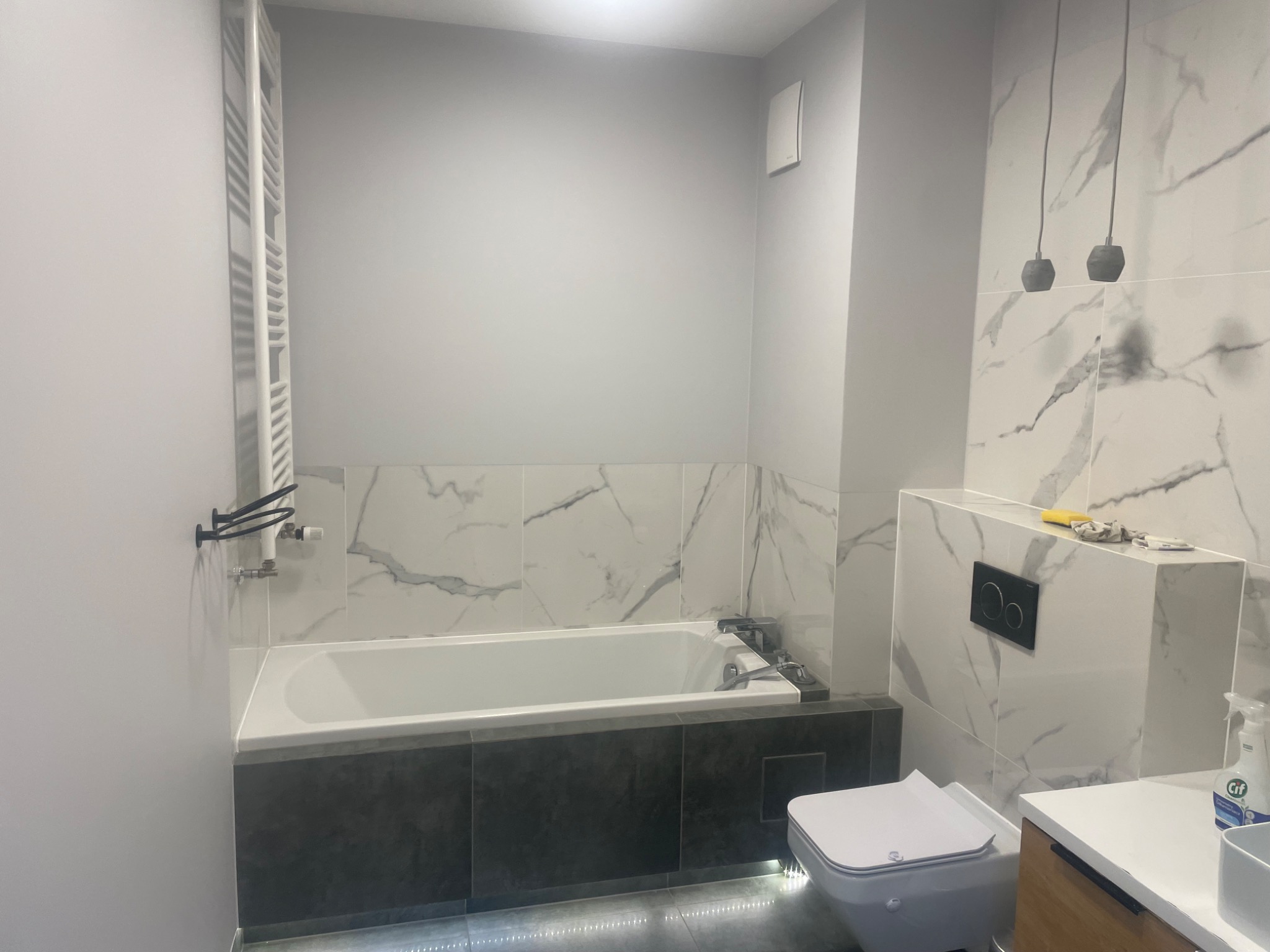 Nowoczesna łazienka po remoncie: biała wanna z szarą obudową, marmurowe płytki, podświetlenie LED pod wanną, wisząca toaleta i minimalistyczne lampy.
