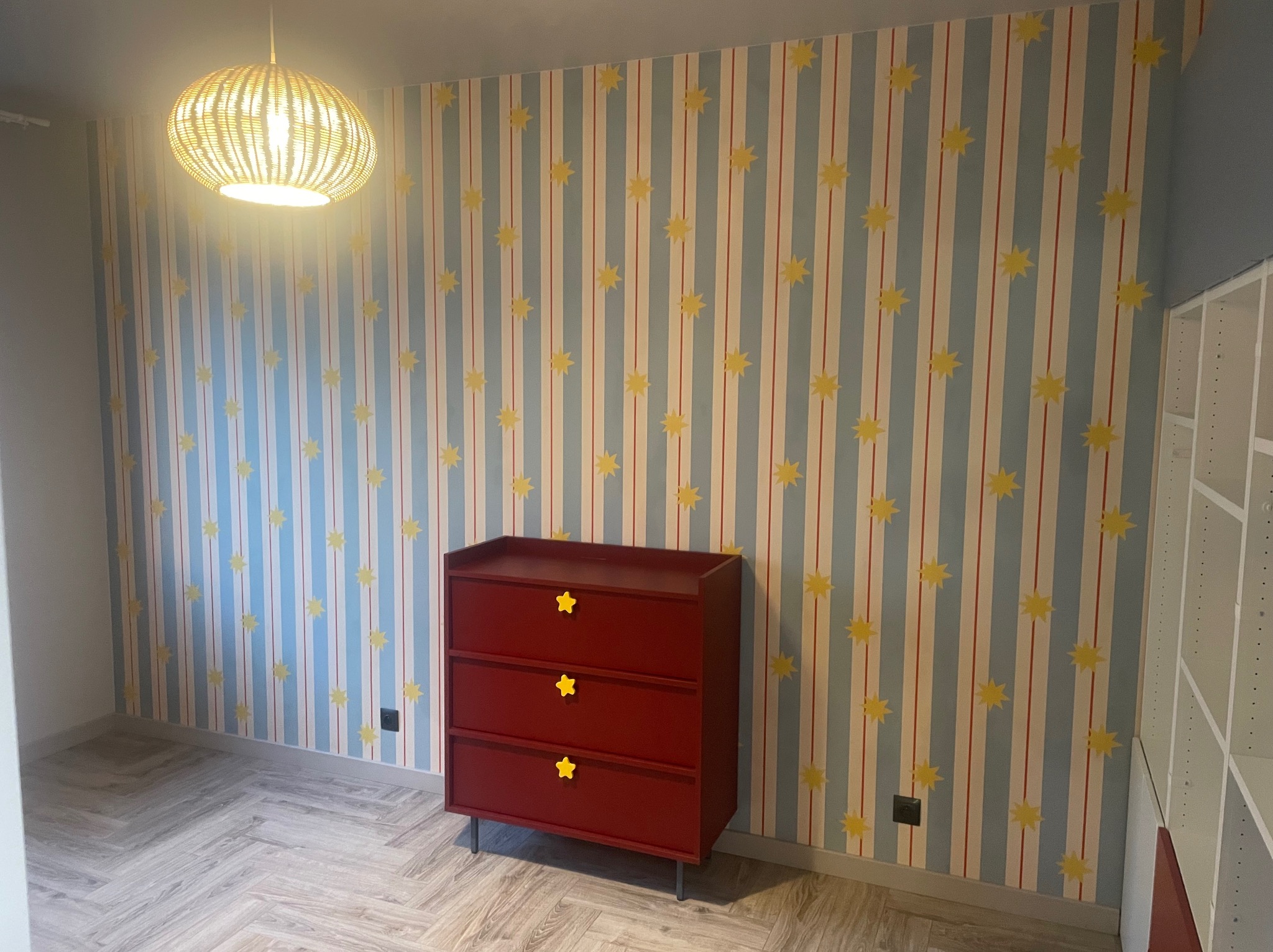 Pokój dziecięcy po odświeżeniu: wzorzysta tapeta w paski i gwiazdki, czerwona komoda z żółtymi uchwytami w kształcie gwiazd, parkiet ułożony w jodełkę, wiklinowa lampa.