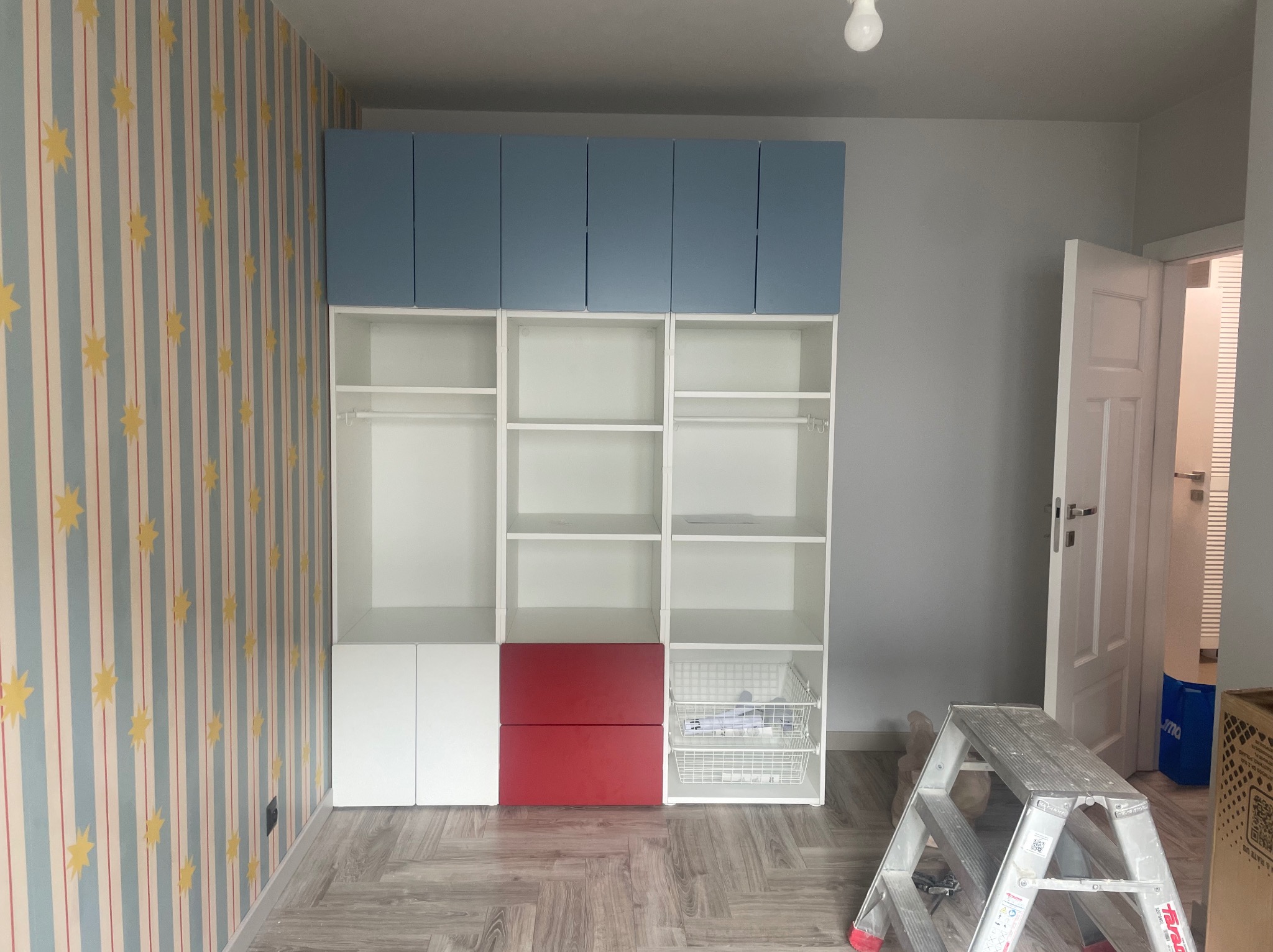 Pokój po remoncie: biało-niebieska szafa z czerwonymi akcentami na tle wzorzystej tapety i szarej ściany. Widoczna drabina malarska.