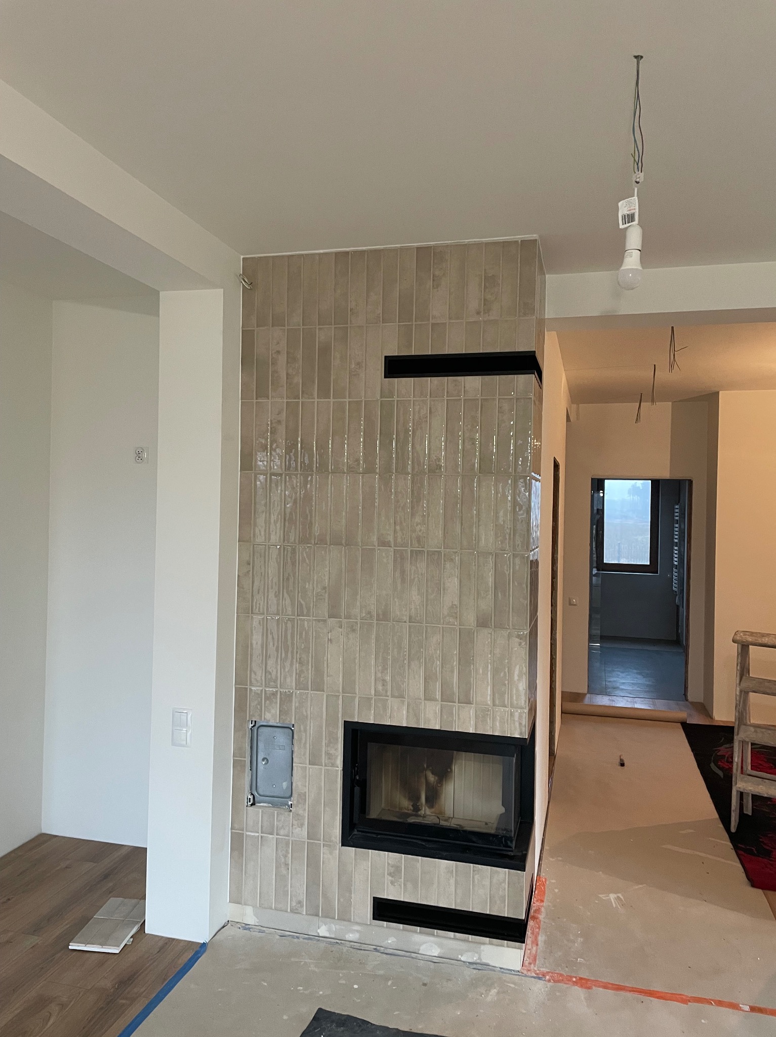 Obudowa kominka z beżowych, błyszczących płytek ułożonych pionowo, z czarnymi akcentami. Widoczne fragmenty podłogi i sufitu w trakcie prac wykończeniowych.