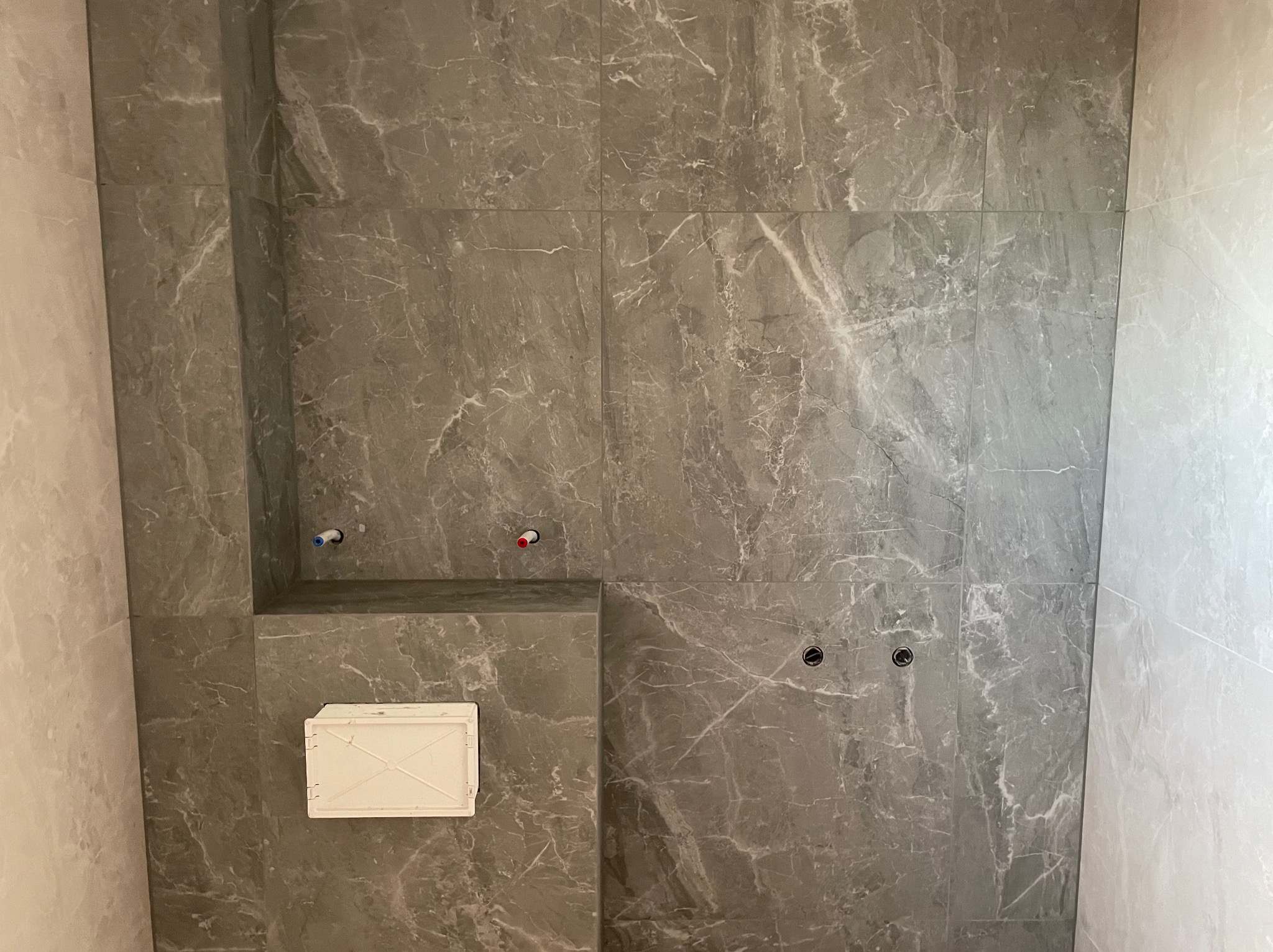 Wykończona ściana łazienki z szarych, marmurkowych płytek, widoczne przyłącza wodne i otwór rewizyjny. Minimalistyczny design, nowoczesne wykończenie.