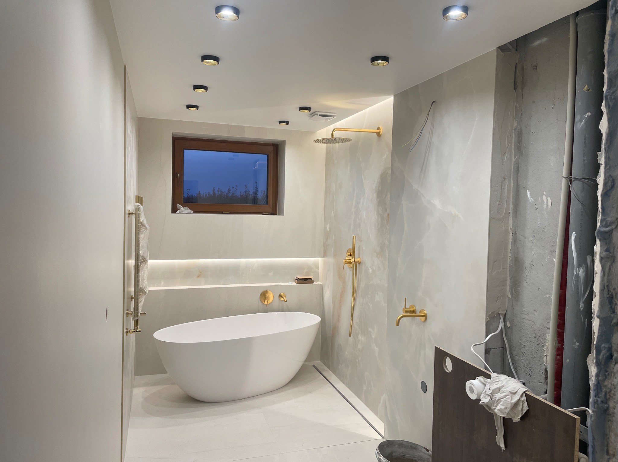 Nowoczesna łazienka w trakcie wykańczania: biała wanna wolnostojąca, złota armatura, podświetlenie LED i fragment ściany w stanie surowym. Widoczne rury i instalacje.