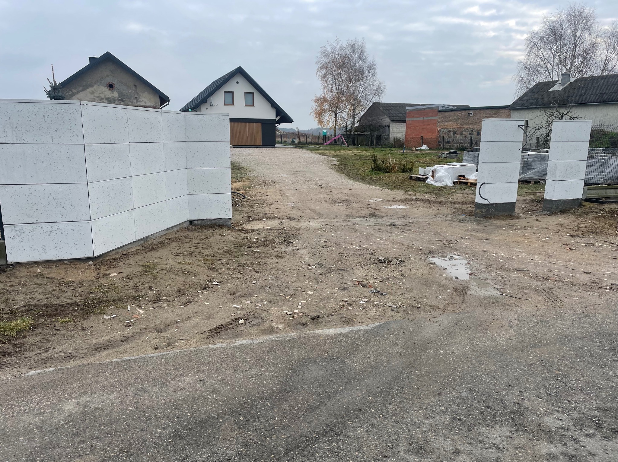 Nowoczesne ogrodzenie modułowe z betonu architektonicznego w trakcie montażu, na tle wiejskiego krajobrazu z domami i polną drogą. Widoczne elementy konstrukcyjne.