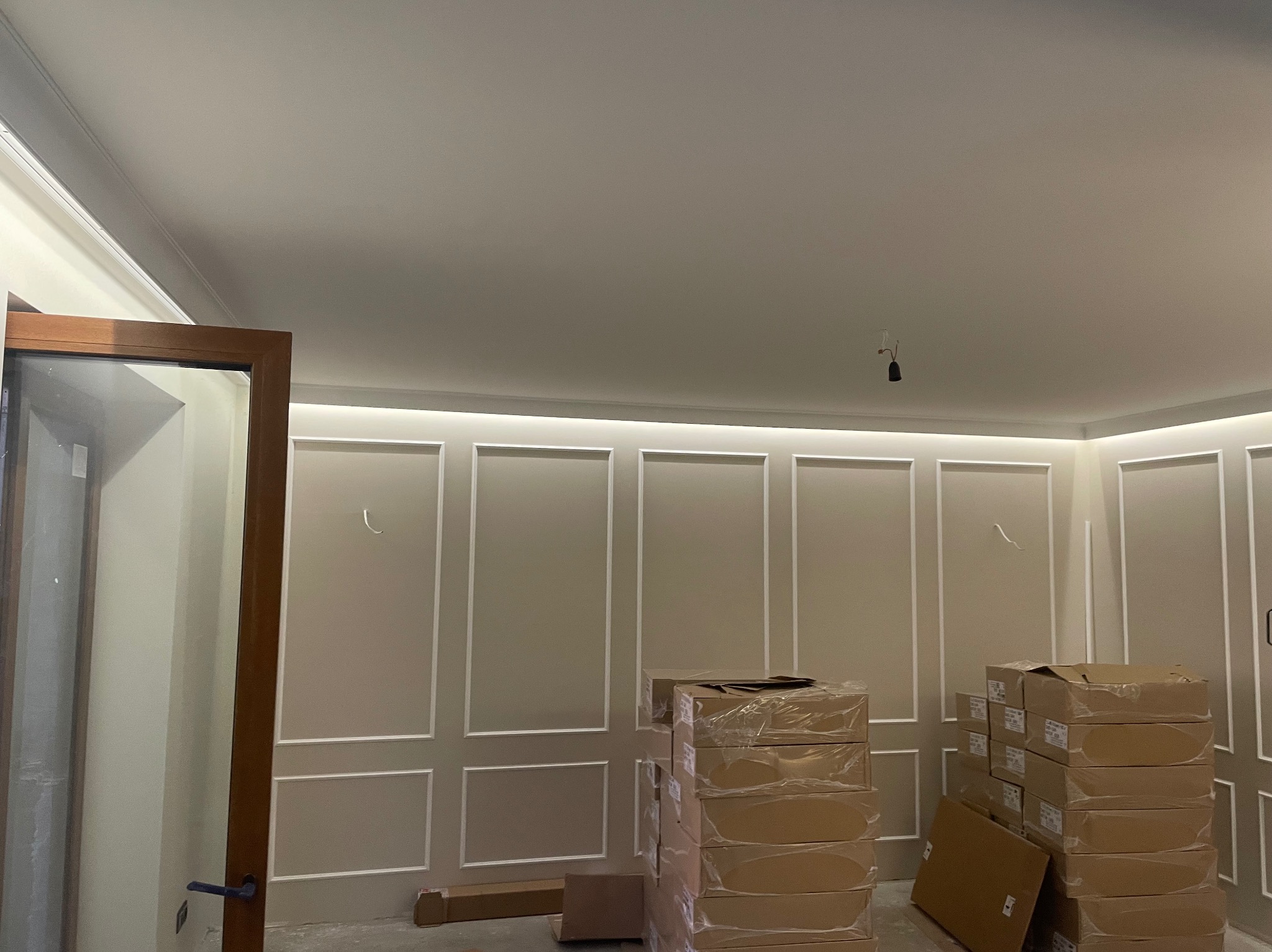 Jasne wnętrze z dekoracyjnymi panelami ściennymi i listwami LED. Stosy zapakowanych materiałów budowlanych czekają na montaż. Widoczna stolarka drzwiowa.