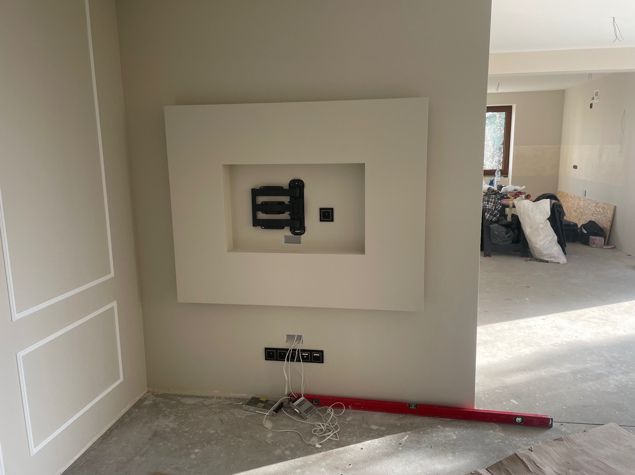 Ściana z zamontowanym uchwytem TV w ozdobnej wnęce, widoczne gniazdka elektryczne i poziomica. W tle surowy stan pomieszczenia w trakcie prac wykończeniowych.