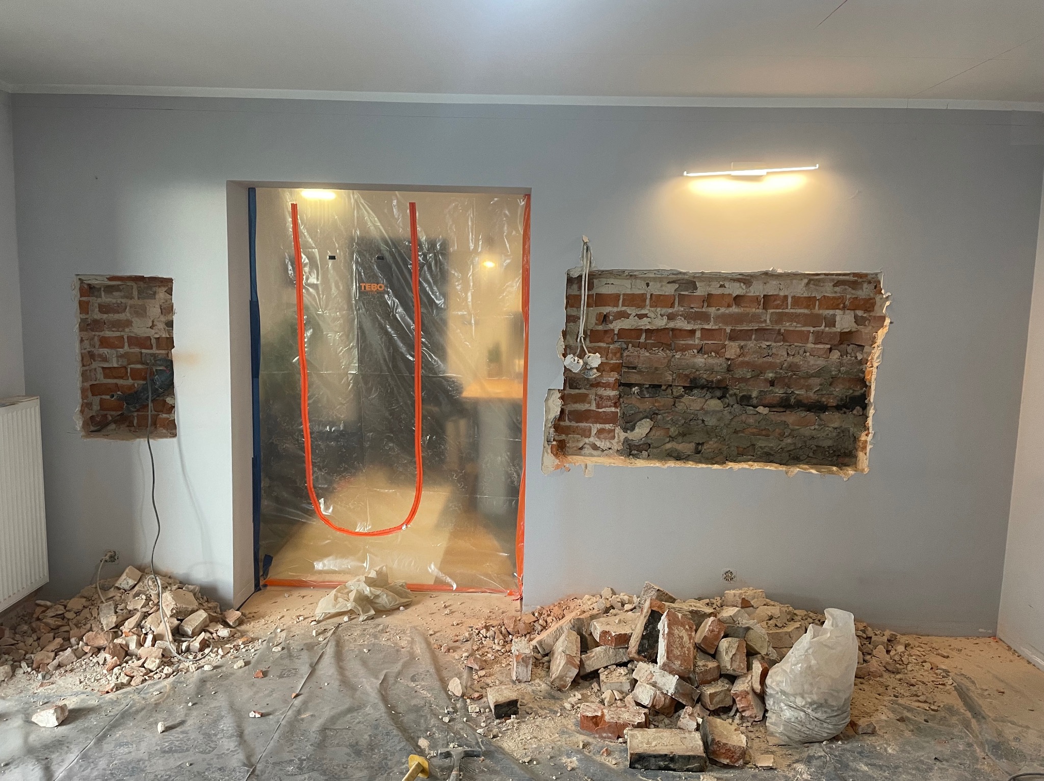 Demontaż fragmentów ściany z cegły w trakcie remontu. Otwór zabezpieczony folią. Gruz na podłodze. Widoczne ślady po kuciu w ścianie. Przygotowanie do dalszych prac wykończeniowych.