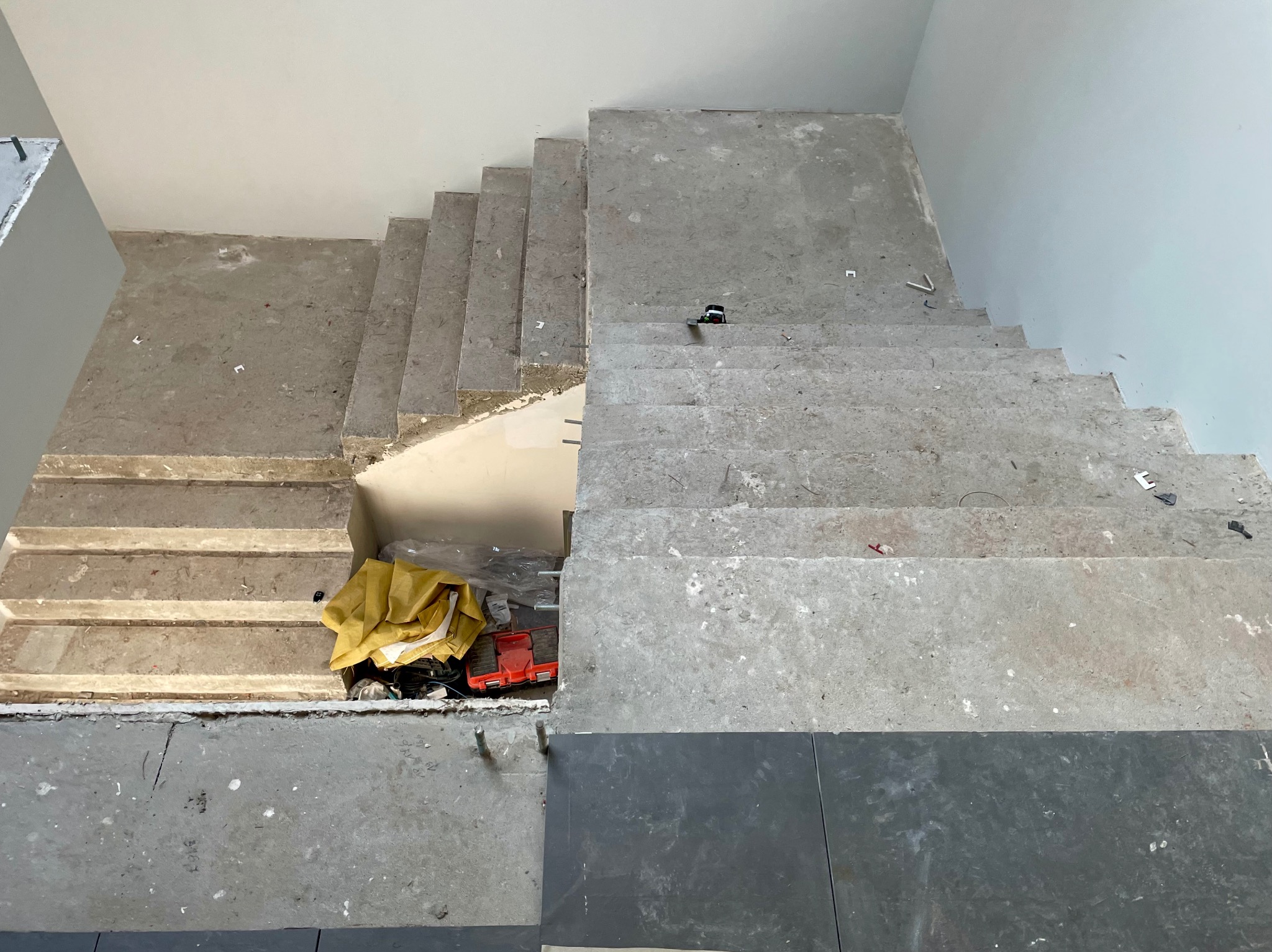 Betonowe schody w trakcie wykańczania, widok z góry. Część schodów z surowego betonu, część z położonymi ciemnymi płytkami. Widoczne narzędzia i materiały budowlane.