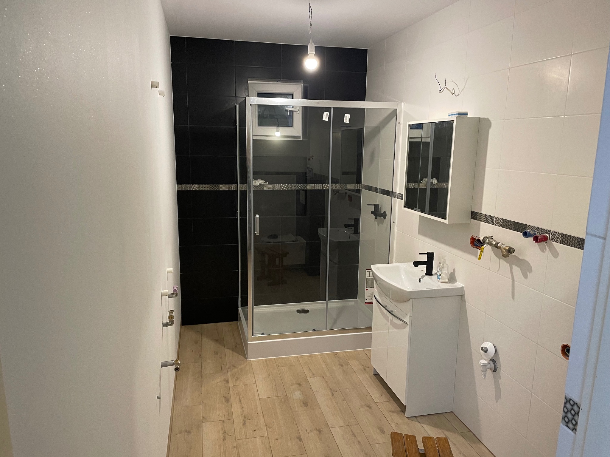 Nowoczesna łazienka po remoncie: kabina prysznicowa ze szklanymi drzwiami, biała umywalka z szafką, czarno-białe płytki, drewnopodobna podłoga i minimalistyczne oświetlenie.