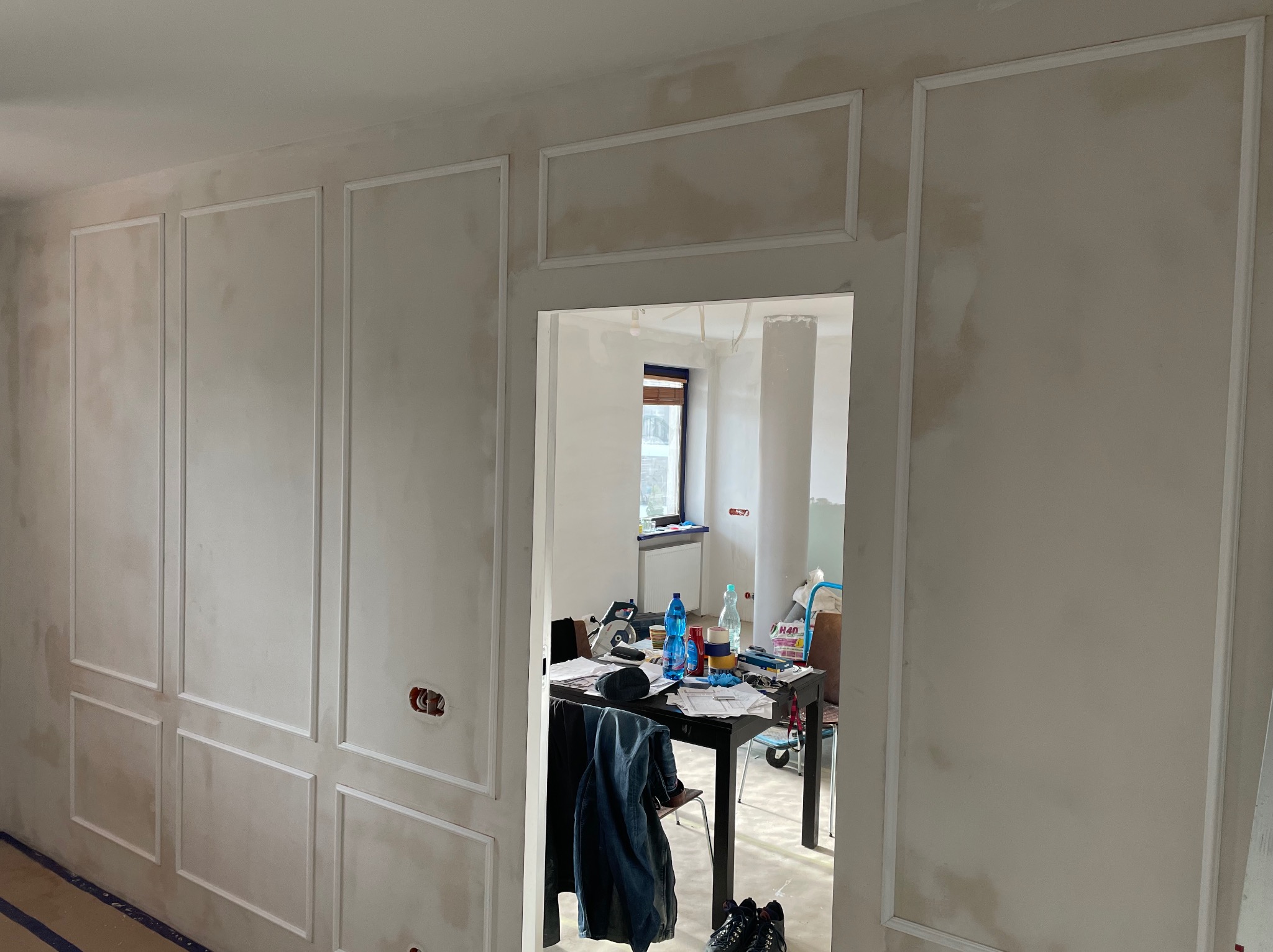 Ściana z dekoracyjnymi listwami w trakcie montażu, widoczne ślady szpachlowania. W tle, przez otwarte drzwi, widoczny stół z narzędziami i materiałami budowlanymi.