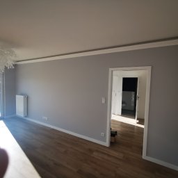 ŁK Wykończenia Wnętrz Łukasz Kłosowski - Jasne wnętrze po malowaniu: szare ściany z białą sztukaterią, drewniana podłoga, widok na balkon i łazienkę. Nowoczesna lampa sufitowa.