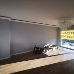 ŁK Wykończenia Wnętrz Łukasz Kłosowski - Wnętrze pokoju po malowaniu ścian na szaro z białymi listwami przypodłogowymi i sufitowymi. Widoczne narzędzia malarskie i podłoga z paneli.