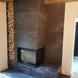 ŁK Wykończenia Wnętrz Łukasz Kłosowski - Wykończony kominek z czarnych płyt w Skokach, z wkładem kominkowym i dekoracyjnym miejscem na drewno. Widoczne drewniane belki stropowe.