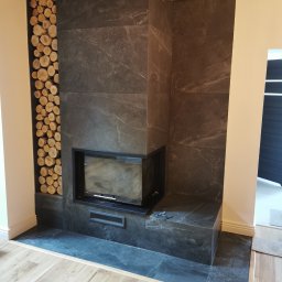 ŁK Wykończenia Wnętrz Łukasz Kłosowski - Obudowa kominka z ciemnego kamienia z wnęką na drewno, wykończona drewnianymi belkami stropowymi i jasną podłogą. Minimalistyczny design.