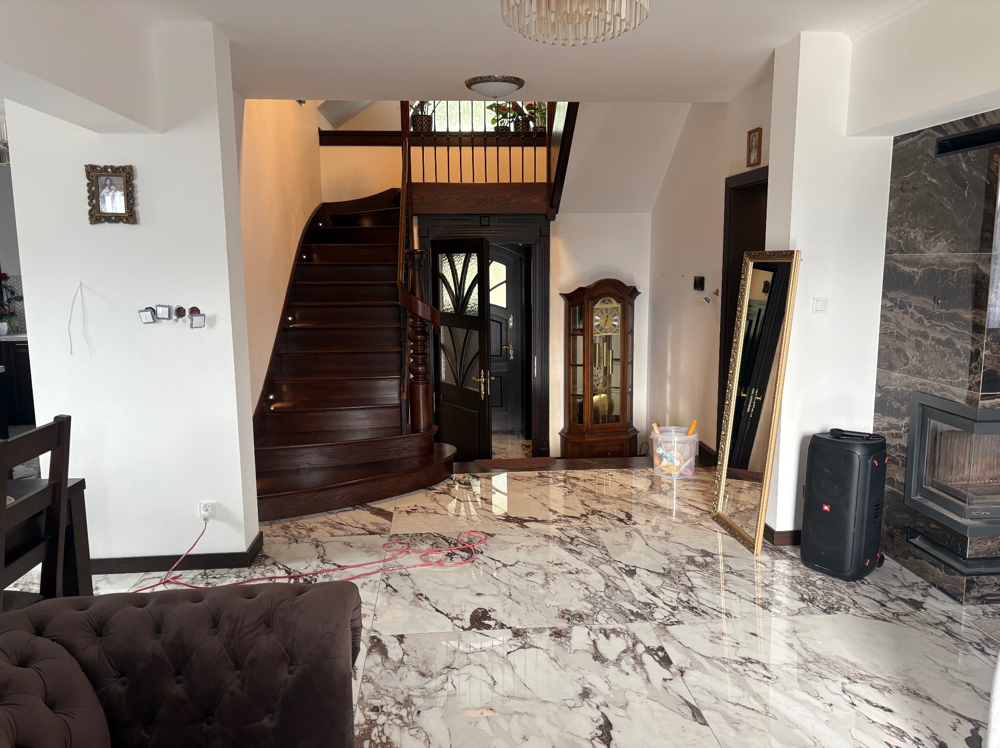 Eleganckie wnętrze domu z marmurową podłogą, drewnianymi schodami i kominkiem z ciemnego kamienia. Widoczne elementy dekoracyjne: lustro w złotej ramie, zegar stojący.