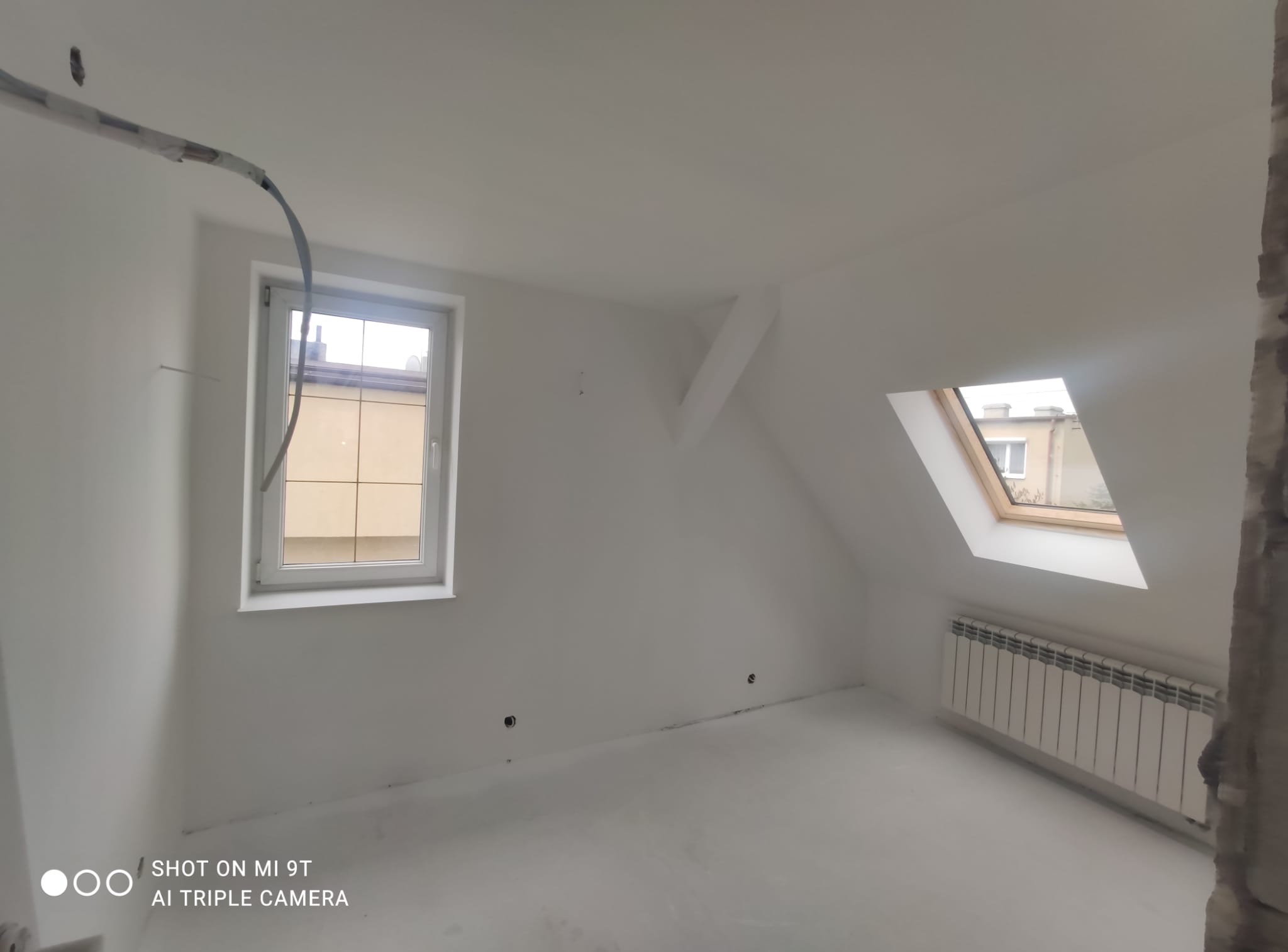 Białe poddasze po remoncie: gładkie ściany, okna dachowe i grzejnik. Widoczne ślady po instalacjach elektrycznych. Minimalistyczny, jasny pokój.