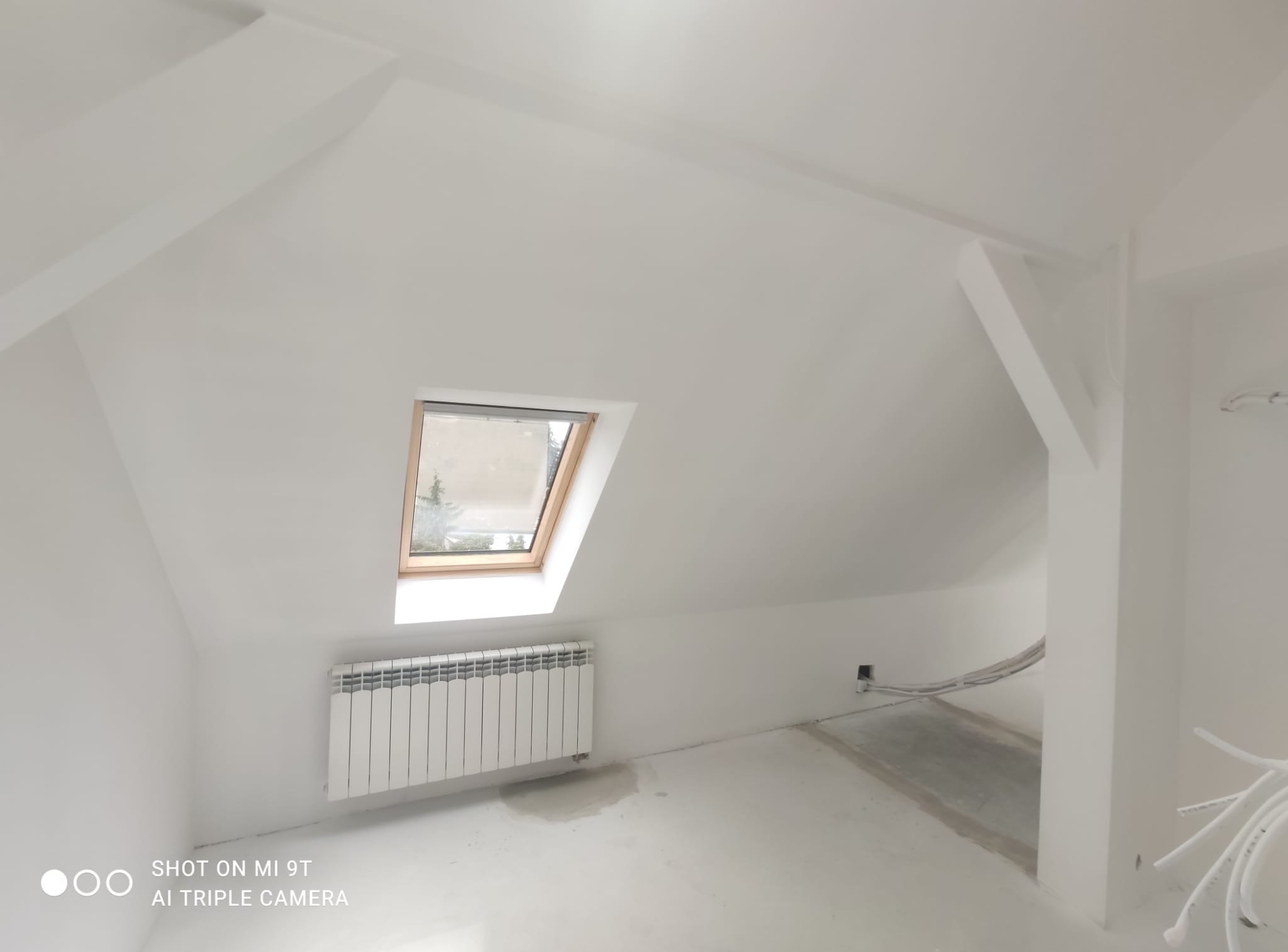 Białe poddasze po remoncie z oknem dachowym, grzejnikiem i widocznymi kablami. Świeżo pomalowane ściany i sufit. Widoczne elementy konstrukcyjne.