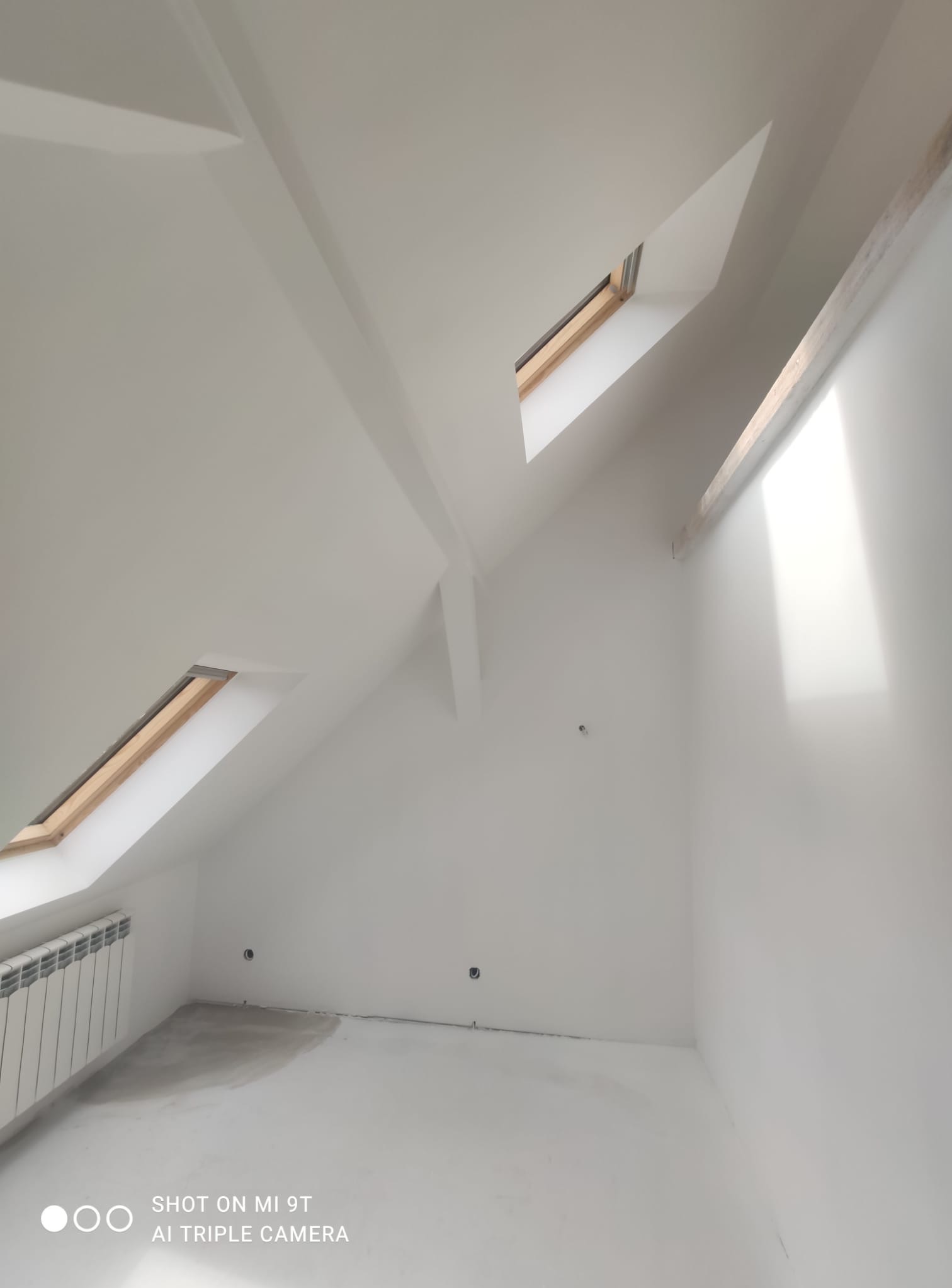 Białe, świeżo pomalowane poddasze ze skosami i oknami dachowymi. Grzejnik przy ścianie, ślady po szpachlowaniu na podłodze. Minimalistyczne wykończenie.