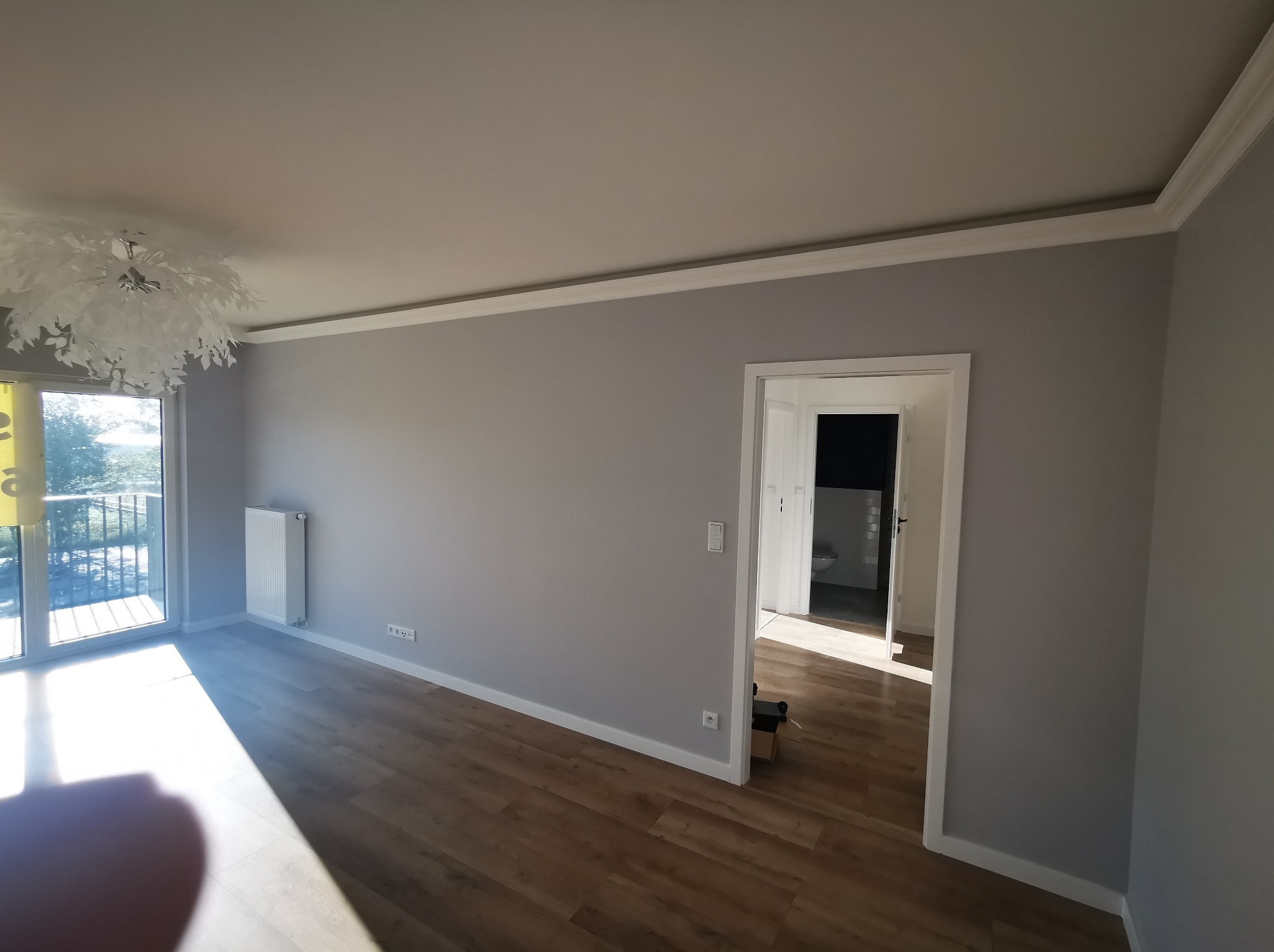 Jasne wnętrze po malowaniu: szare ściany z białą sztukaterią, drewniana podłoga, widok na balkon i łazienkę. Nowoczesna lampa sufitowa.