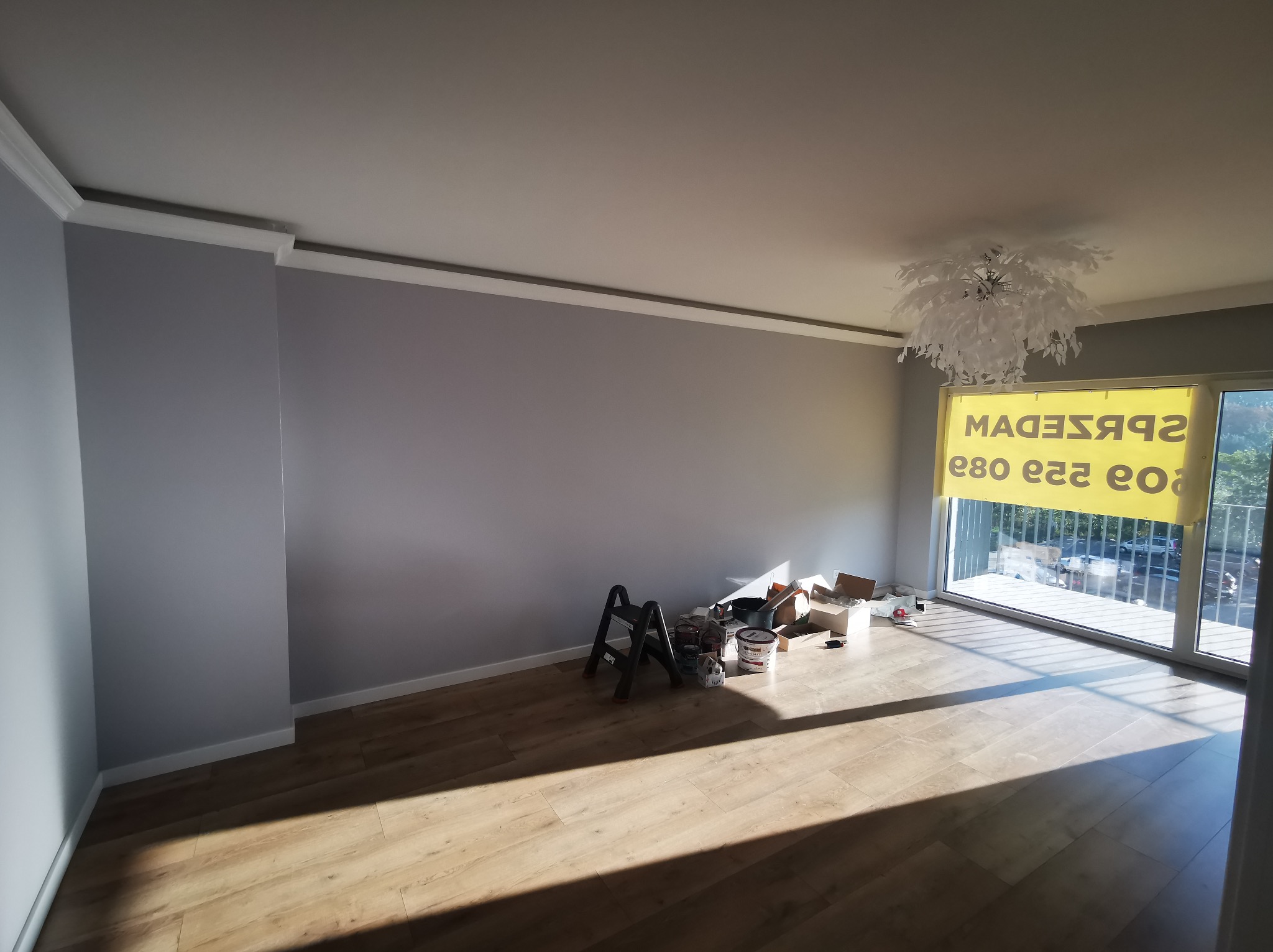 Wnętrze pokoju po malowaniu ścian na szaro z białymi listwami przypodłogowymi i sufitowymi. Widoczne narzędzia malarskie i podłoga z paneli.