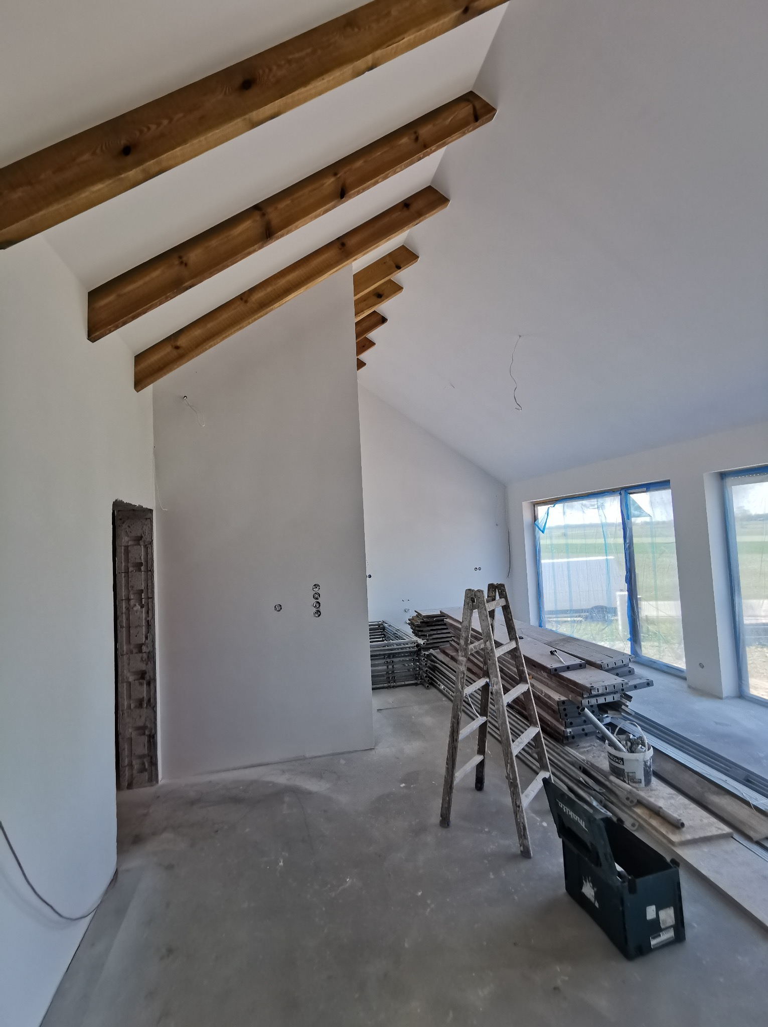 Wnętrze w trakcie remontu: białe ściany, drewniane belki stropowe, drabina, narzędzia i materiały budowlane. Widoczne przygotowania do dalszych prac wykończeniowych.
