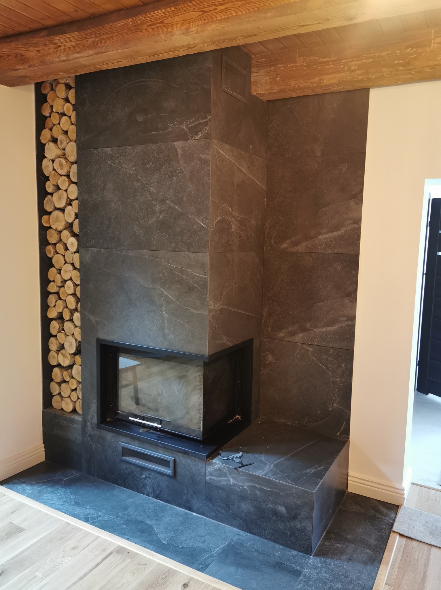 Wykończony kominek z czarnych płyt w Skokach, z wkładem kominkowym i dekoracyjnym miejscem na drewno. Widoczne drewniane belki stropowe.