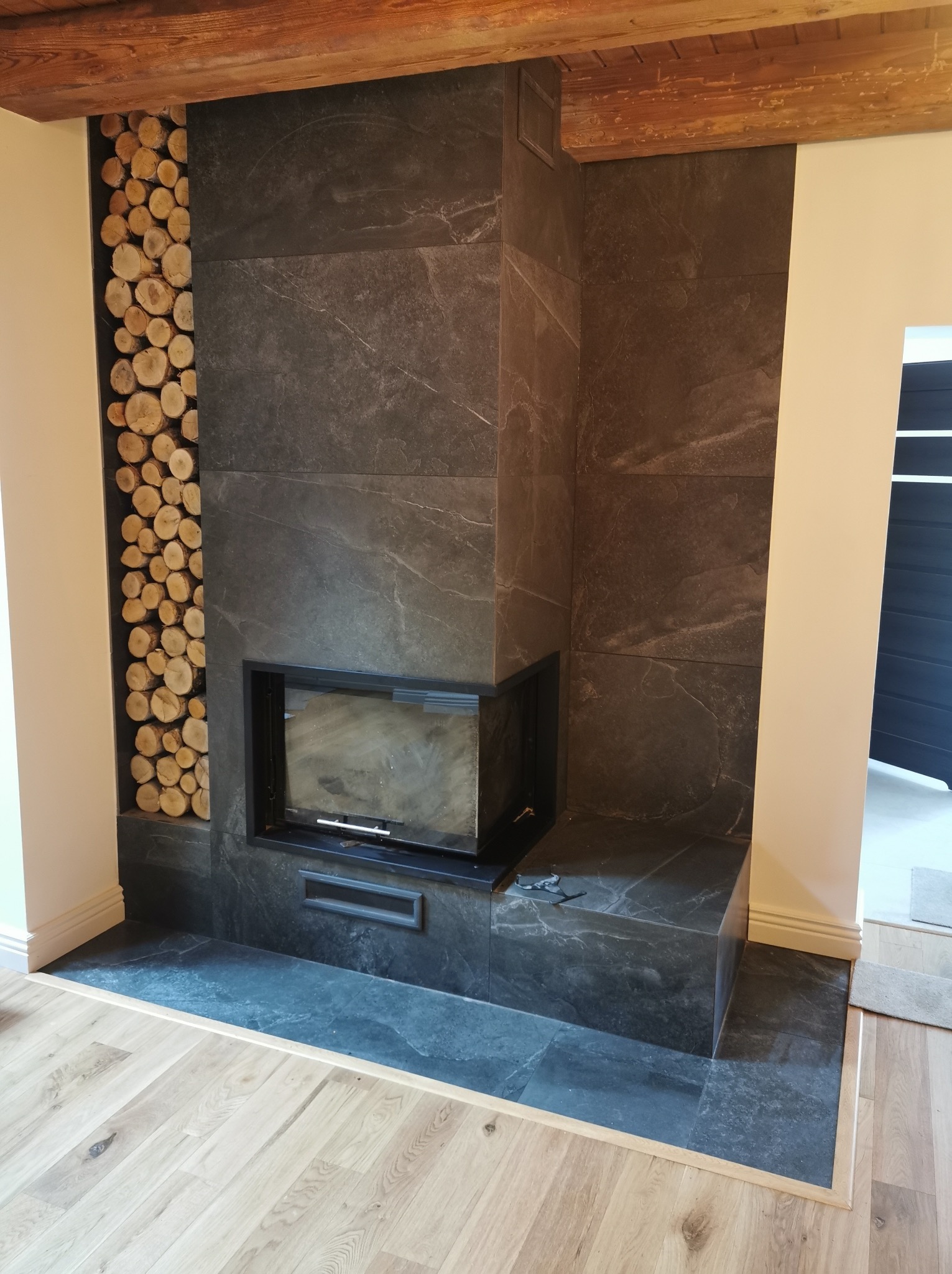 Obudowa kominka z ciemnego kamienia z wnęką na drewno, wykończona drewnianymi belkami stropowymi i jasną podłogą. Minimalistyczny design.