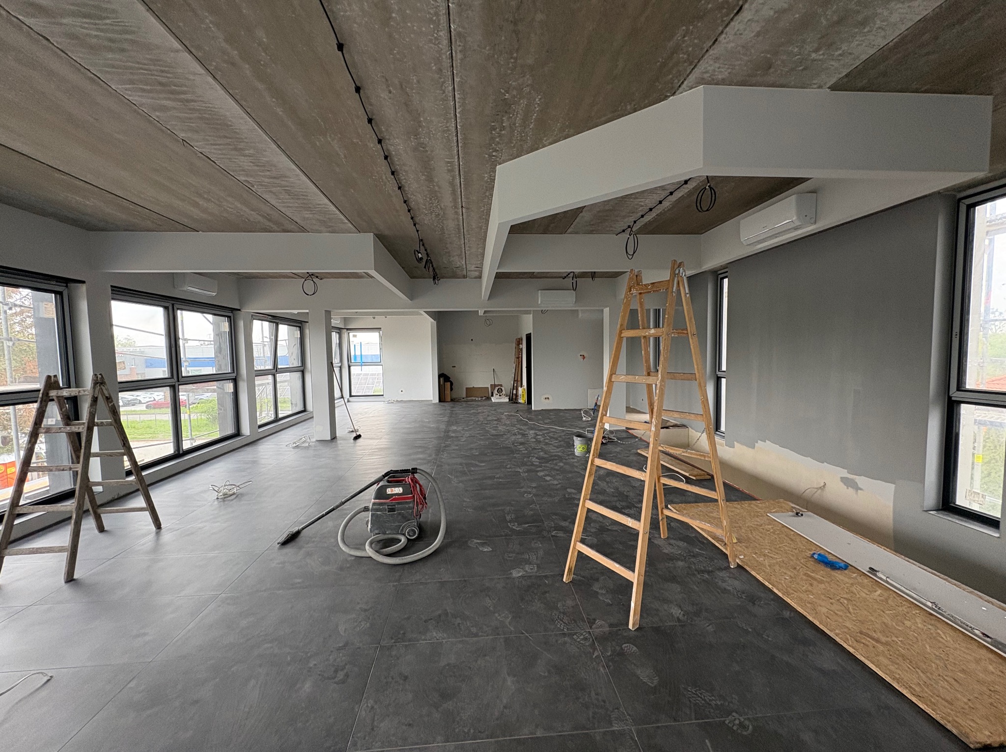 Wnętrze w trakcie prac wykończeniowych: szare płytki podłogowe, surowy betonowy sufit, drabiny, odkurzacz przemysłowy i częściowo pomalowane ściany na szaro.