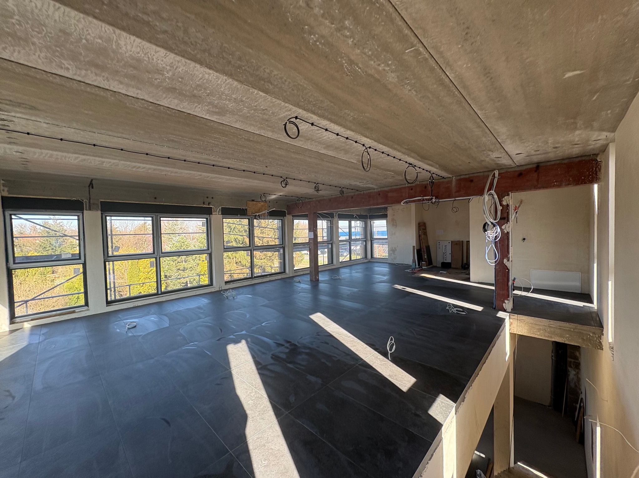 Wnętrze w trakcie remontu: surowy betonowy strop z instalacją elektryczną, duże okna z widokiem na zieleń, ciemna podłoga z płytek. Widoczne elementy konstrukcyjne i schody.