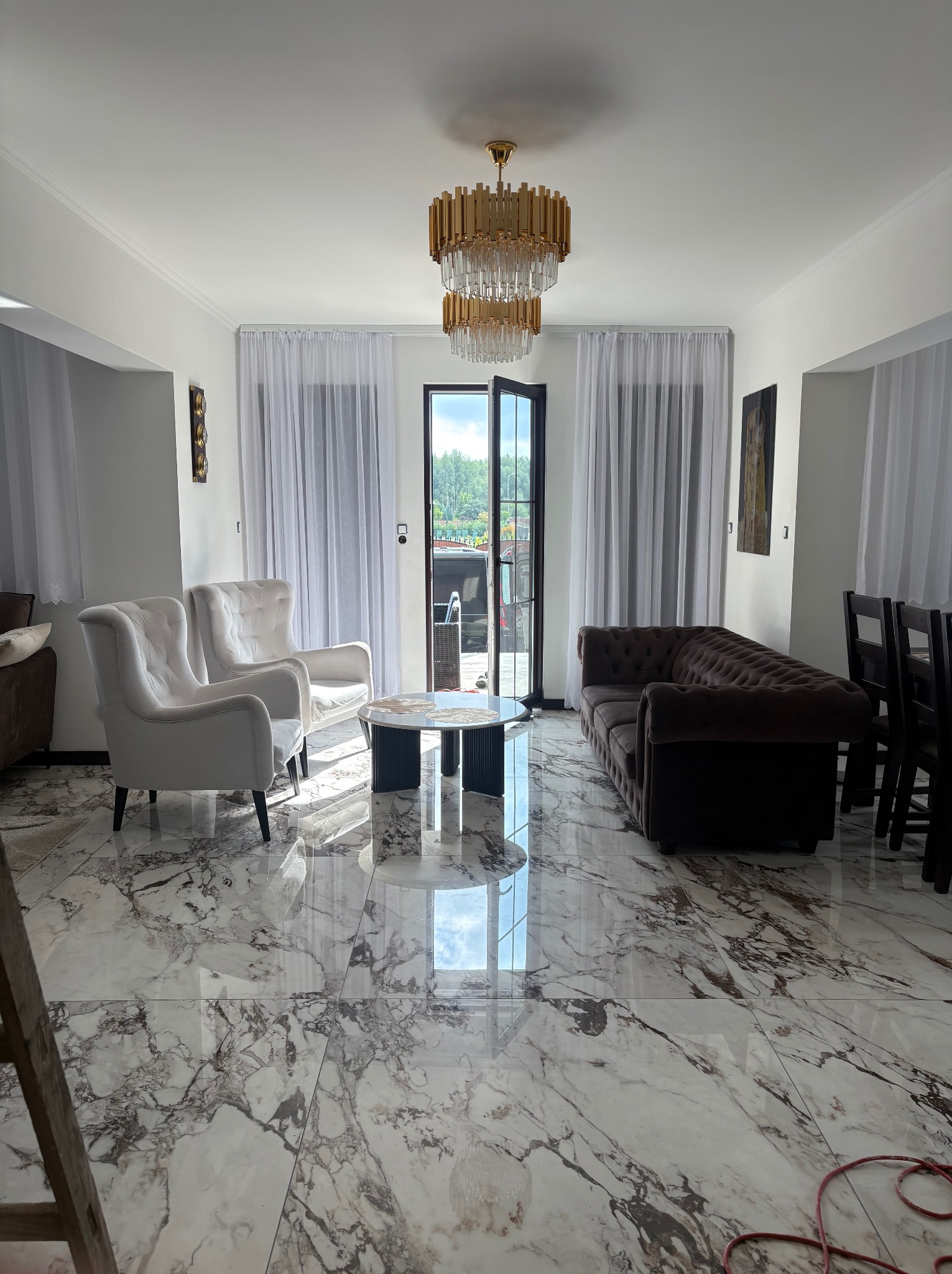 Elegancki salon z marmurową podłogą, kremowymi fotelami i brązową sofą. Centralnie umieszczony żyrandol dodaje luksusowego charakteru. Widoczne drzwi balkonowe z białymi zasłonami.