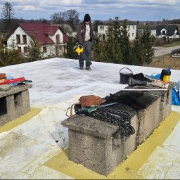 Jakub Turzyński - Biały dach pokryty pianą montażową, z kominami i narzędziami. W tle pracownik z dmuchawą i zabudowa jednorodzinna. Widok z góry, szeroki kadr.