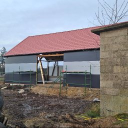 Jakub Turzyński - Budynek z czerwonym dachem w trakcie ocieplania elewacji, widoczne rusztowania i przygotowane materiały. Stara stodoła z drewnianymi wrotami w tle.