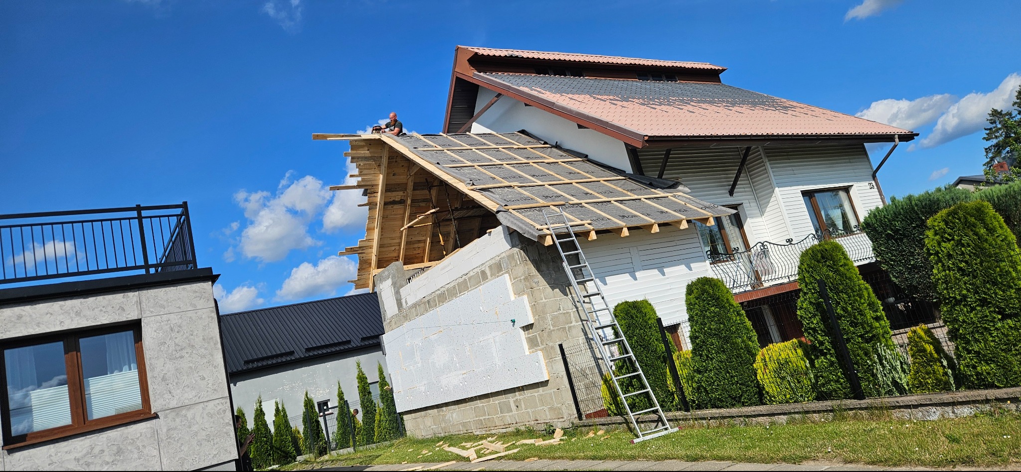 Dom w trakcie wymiany dachu, widoczny dekarz pracujący na konstrukcji. Część dachu pokryta nową membraną i łatami, obok drabina oparta o ścianę budynku z ociepleniem.