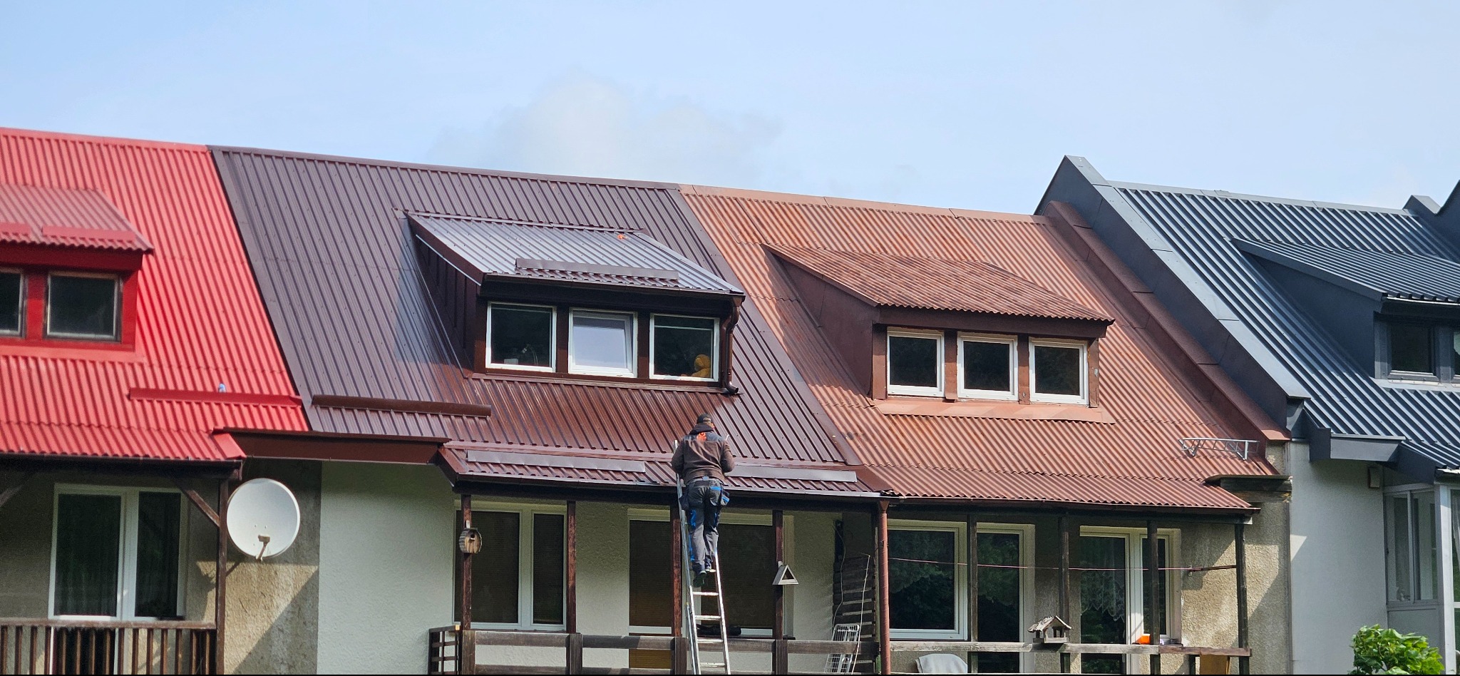 Różnokolorowe dachy z blachy falistej na szeregu domów. Dekarz na drabinie przy brązowym dachu. Widoczne okna dachowe i elewacja z balkonami.