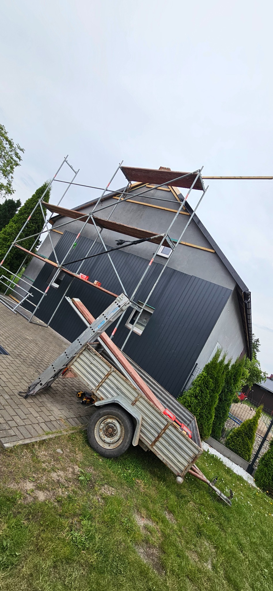 Dom z szarą elewacją w trakcie prac dekarskich, widoczne rusztowania i przyczepka z materiałami. Ujęcie z dołu, pochmurne niebo.