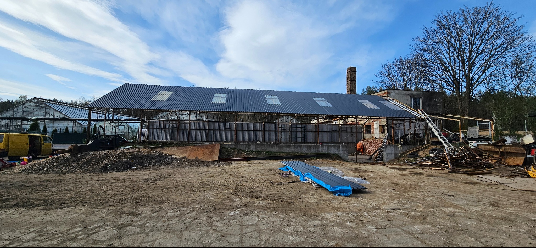 Nowy, ciemny dach z świetlikami na konstrukcji budynku gospodarczego. Na ziemi leżą arkusze blachy. W tle szklarnia i drzewa pod błękitnym niebem.